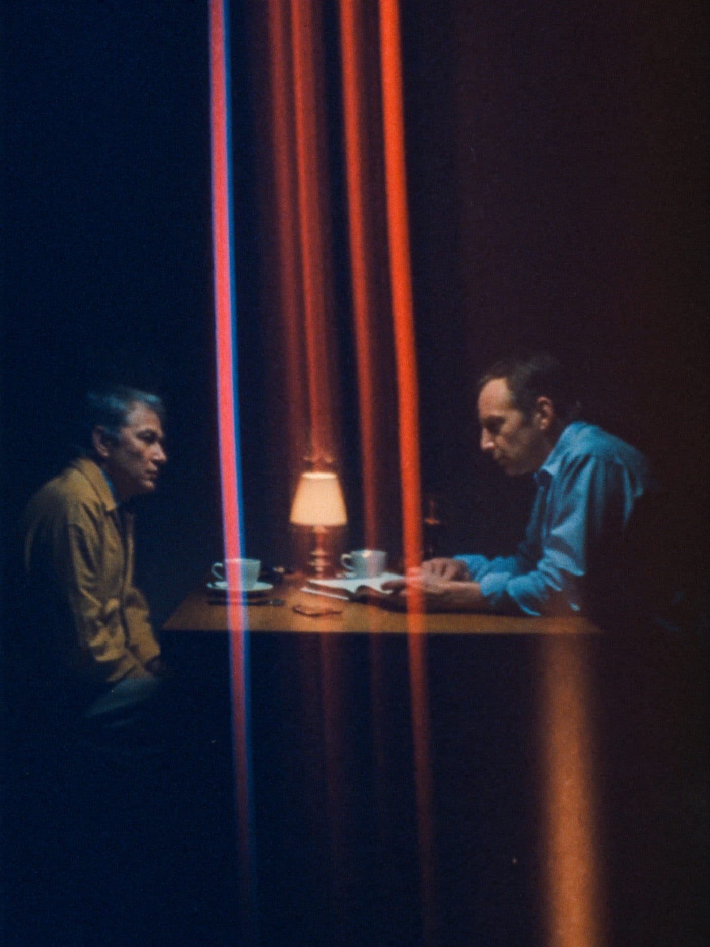 Twee mannen in gesprek aan een kleine tafel met koffie, het licht van een lamp tussen hen.
